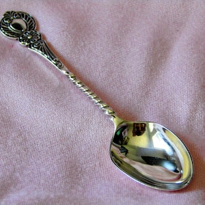 May include: A silver-toned metal spoon featuring a twisted handle and an ornate, decorative finial at the top. The handle transitions into a smooth, polished oval bowl. The metal surface exhibits a reflective finish with intricate embossed details.