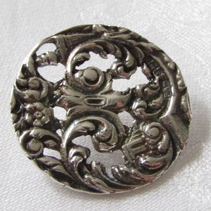 May include: A circular silver-toned metal button featuring an intricate openwork design of swirling acanthus leaves and floral motifs. The surface has a polished, reflective finish with recessed areas that highlight the detailed relief patterns.