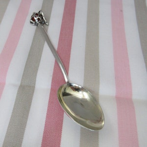 May include: A silver spoon with a decorative handle in the shape of a bird. The spoon is lying on a striped pink and gray fabric.