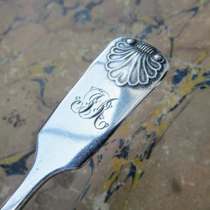 Puede incluir: Primer plano de una cuchara de plata con un diseño decorativo de concha en la parte superior. La cuchara presenta un monograma grabado de dos letras entrelazadas. El mango de la cuchara es liso y pulido.