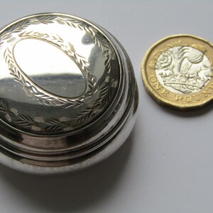Puede incluir: Un pequeño recipiente redondo plateado con un diseño ovalado en la tapa, rodeado por un patrón de hojas decorativas. Una moneda de una libra está al lado del recipiente.