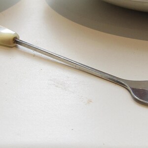 May include: A small silver-toned metal cocktail fork featuring a slender, straight handle and a three-pronged head. The end of the handle is capped with a smooth, rectangular, off-white pearlescent material that provides a decorative accent.