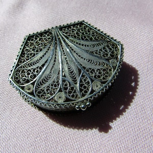 May include: A silver filigree box with a fan-shaped design on the lid. The box has an intricate, openwork pattern and a scalloped edge. The box is set against a light pink background.