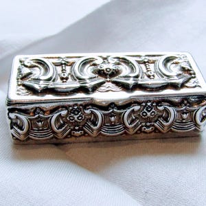 May include: A rectangular silver-toned metal box featuring intricate embossed scrollwork and floral patterns on the lid and sides. The surface has a polished, reflective finish with deep grooves that highlight the detailed relief designs.