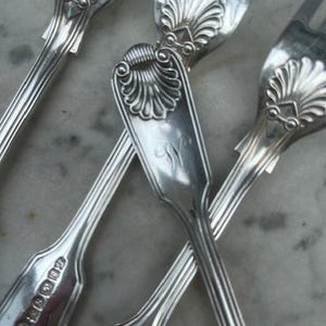May include: Close-up of several antique silver forks. The forks have a decorative shell design on the handle and tines. The silver forks are arranged in a crisscross pattern against a marble surface.