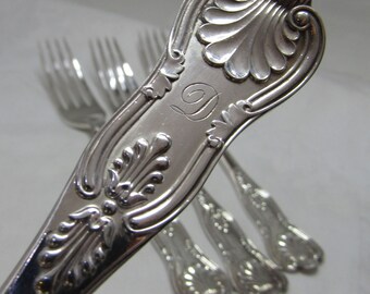 May include: Close-up of a silver fork with ornate, engraved floral and leaf designs. The handle features a stylized 'D' monogram. Several other forks are slightly out of focus in the background, suggesting a set of silverware.