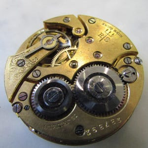 May include: Close-up of a gold-toned pocket watch movement with intricate gears and a trident logo. The watch is labelled "Swiss Made" and "RIS".