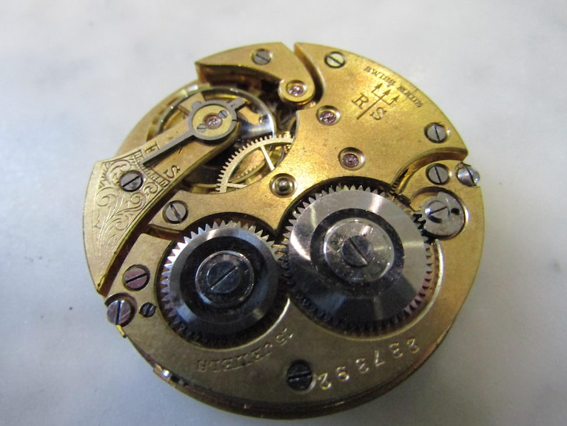 May include: Close-up of a gold-toned pocket watch movement with intricate gears and a trident logo. The watch is labelled "Swiss Made" and "RIS".