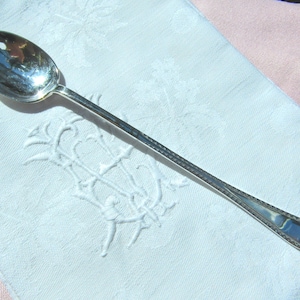 May include: A silver serving fork with a three-pronged head, on a white embroidered cloth. The fork has a long, slender handle with a decorative pattern. The cloth is on a pink surface.
