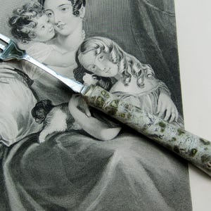 May include: A silver fork with three prongs and a handle featuring a mottled green and white design. The fork rests on a black and white print of a woman and children.