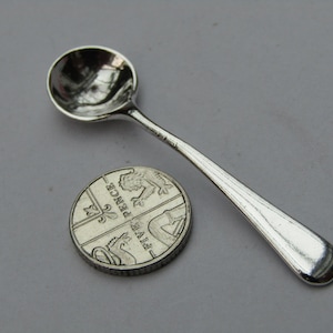 Puede incluir: Una pequeña cuchara plateada con un cuenco redondeado y un mango largo, junto a una moneda de cinco peniques. La cuchara y la moneda están sobre una superficie gris. La cuchara probablemente se utiliza para servir porciones pequeñas.