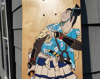 May include: A wooden skateboard deck with a hand-painted design of a samurai warrior in blue, yellow, and black clothing holding a sword. The background is a light gold with a white cloud.