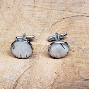 May include: A pair of silver cufflinks with oval-shaped, clear, white, and black speckled stones. The cufflinks are set in a silver metal frame and have a classic T-bar closure. The background is a light brown wooden surface.