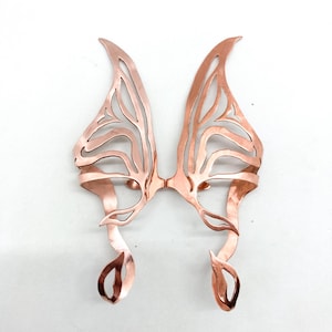 May include: Copper-colored, open-work ear cuffs in a butterfly wing design. The symmetrical pair features intricate cut-out patterns and curved edges, creating a delicate, nature-inspired accessory. The ear cuffs are designed to be worn on the ears.