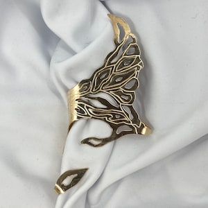 May include: A gold-colored napkin ring with an openwork design. The ring has a band that wraps around the napkin and a decorative, leaf-like element. The design is intricate and detailed, adding a touch of elegance to the table setting.