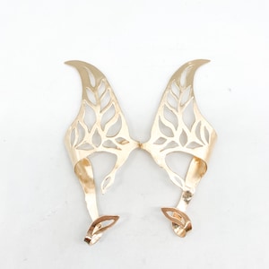 May include: Gold-colored ear cuffs with a leaf-like design. The cuffs have a curved shape that wraps around the ear, with intricate cut-out patterns resembling leaves. The ear cuffs are made of a shiny metal and are designed to be worn on the ears.