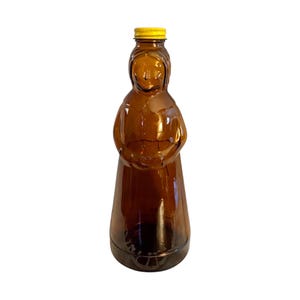 May include: A translucent amber-colored glass bottle shaped like a person wearing a bonnet and a long dress. The bottle features a molded facial profile and a flared base, topped with a small, ribbed, bright yellow screw-on plastic cap.