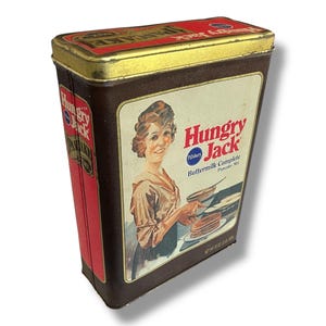 May include: Vintage Hungry Jack Buttermilk Complete Pancake Mix tin. The rectangular tin has a brown body with a gold-colored lid. The front features a woman and the words "Hungry Jack" and "Pillsbury Buttermilk Complete Pancake Mix."