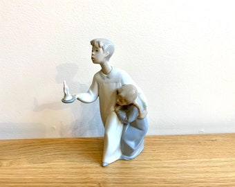 May include: A porcelain figurine of a boy holding a candle and carrying a smaller boy on his back. The figurine is painted in shades of white and blue.