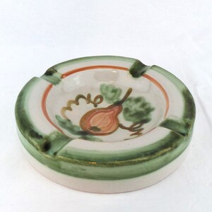 Puede incluir: Un cenicero de cerámica blanca con un borde verde y naranja. El cenicero tiene un diseño pintado de una pera con hojas.