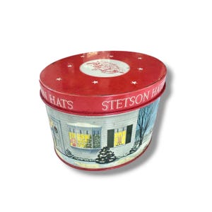 Könnte beinhalten: Eine Vintage-Stetson-Hüte-Dose mit rotem Deckel, der eine weiße Illustration und Sterne zeigt. Die zylindrische Dose hat eine Winterszene mit einem Haus und einem Weihnachtsbaum. Die Worte "STETSON HATS" sind aufgedruckt.