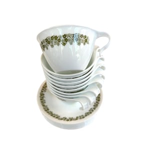 May include: A stack of white ceramic cups and saucers featuring a repeating olive green floral pattern around the rims. The cups have curved handles and are nested together, while the matching saucers are stacked neatly at the base of the set.