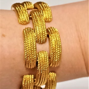 May include: A gold-toned bracelet with a textured, chain-link design. The bracelet is made of a shiny, metallic material and is worn on a wrist. The links are rectangular and have a ridged surface, creating a detailed and eye-catching accessory.