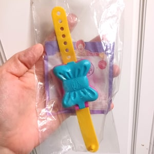 May include: A yellow and blue plastic toy watch, still in its original packaging. The watch features a yellow strap with holes and a blue chef hat design. The packaging has a purple border and a Ronald McDonald image.