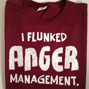 May include: A maroon t-shirt with the text "I Flunked Anger Management" in white, distressed font. The shirt is folded neatly, showcasing the bold statement across the front. A simple, casual garment.