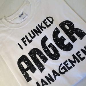 May include: White t-shirt with the text "I Flunked Anger Management" in large, distressed black letters. The shirt is made of a soft, cotton material and is folded neatly. A Gildan label is visible.