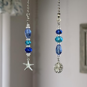 May include: Two beaded fan pull chains with ocean-themed charms. One chain features a starfish charm, while the other has a sand dollar charm. The beads are shades of blue and turquoise, with silver-tone chain.