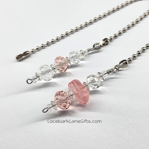 May include: Two beaded ceiling fan pull chains. Each chain features clear and pink faceted beads, with a larger pink bead in the center. The chains are attached to a silver-colored ball chain.