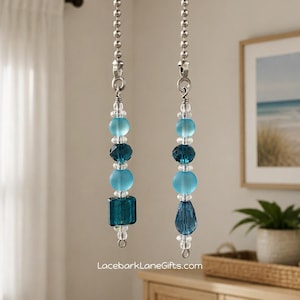 May include: Two ceiling fan pull chains featuring silver-toned ball chains attached to decorative beaded ornaments. Each ornament consists of clear, frosted light blue, and deep teal glass beads in various shapes, including spheres, a square, and a teardrop.