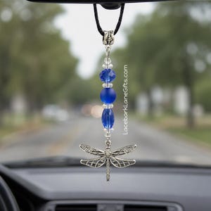 May include: A hanging car ornament featuring a silver-toned dragonfly charm suspended from a black cord. The design includes a vertical arrangement of blue faceted beads, a matte blue round bead, and clear spacer beads. Text reads LacebarkLaneGifts.com.