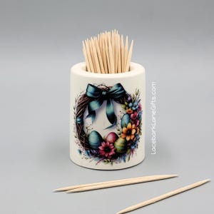 May include: A white ceramic toothpick holder with an Easter wreath design. The wreath features a teal bow, colorful eggs, and flowers. The holder is filled with wooden toothpicks. The text "LacebarkLaneGifts.com" is visible on the side.