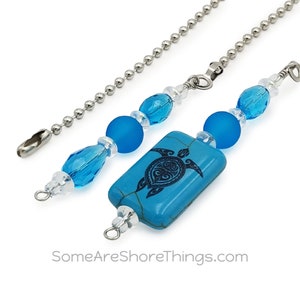 May include: Decorative ceiling fan pull chains with a turquoise turtle pendant. The chains feature blue and clear beads, with a rectangular turquoise pendant showcasing a black turtle design. The chains are made of silver-colored metal.