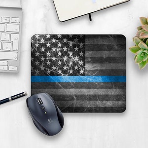 May include: A rectangular mousepad featuring a distressed American flag design in shades of gray and black, with a vibrant blue stripe. A wireless mouse sits below the mousepad. A keyboard, pen, and notebook are also visible.