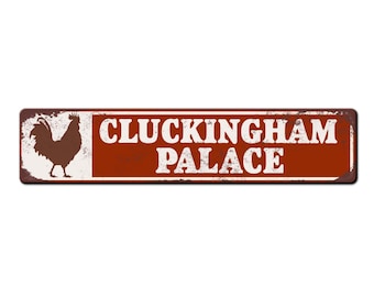 May include: A rectangular, rusty-red metal sign with the words "CLUCKINGHAM PALACE" in white, distressed lettering. A brown rooster silhouette is on the left. The sign has a vintage, weathered appearance.