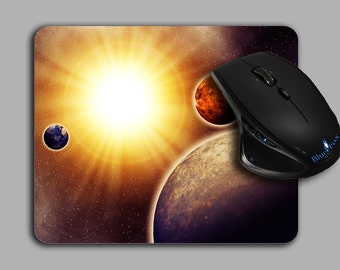 May include: A black computer mouse with a blue logo that says "BlueFox" on the side. The mouse is positioned on a space-themed mousepad with a bright yellow sun, a red planet, and a blue planet.