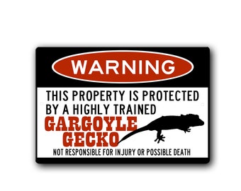 May include: A black and red warning sign with the text "WARNING" in a red oval. The text "THIS PROPERTY IS PROTECTED BY A HIGHLY TRAINED GARGOYLE GECKO" is in white. The text "NOT RESPONSIBLE FOR INJURY OR POSSIBLE DEATH" is in white. A black silhouette of a gecko is shown.