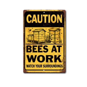 May include: A rectangular metal sign with a yellow background and black text reading CAUTION BEES AT WORK WATCH YOUR SURROUNDINGS. The design features illustrations of two wooden beehives and flying bees, with a distressed, weathered texture.