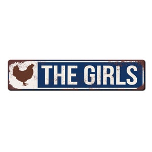 Puede incluir: Un letrero de metal azul y blanco con textura oxidada. El letrero presenta una silueta de un pollo y el texto "THE GIRLS".