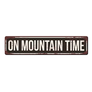 May include: A rusty metal sign with black background and white text that reads "ON MOUNTAIN TIME".