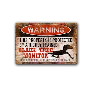 May include: A rectangular metal sign with a distressed, rusted texture. It features a red oval at the top with the white text WARNING. Below, black and red text reads: THIS PROPERTY IS PROTECTED BY A HIGHLY TRAINED BLACK TREE MONITOR NOT RESPONSIBLE FOR INJURY OR POSSIBLE DEATH. A black silhouette of a lizard is centered on the right side.