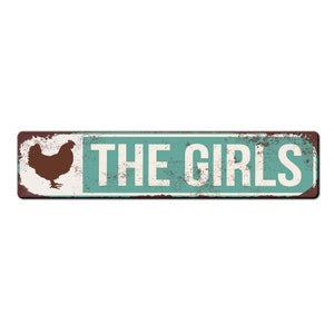 Puede incluir: Un letrero de metal r&uacute;stico con un fondo azul verdoso desgastado y letras blancas. El letrero presenta una silueta de un pollo y el texto "THE GIRLS".
