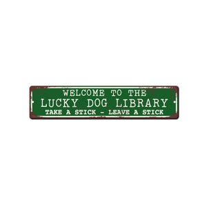 May include: A rectangular, green metal sign with white text that reads "WELCOME TO THE LUCKY DOG LIBRARY" and "TAKE A STICK - LEAVE A STICK." The sign has a distressed, rusty brown border.