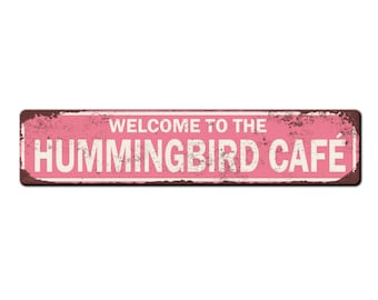 Può includere: Un cartello in metallo di stile vintage rosa e marrone con il testo "Benvenuti al Hummingbird Cafe".