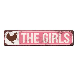 Puede incluir: Un letrero de metal rosa con una silueta de pollo marr&oacute;n y el texto "THE GIRLS" en blanco.