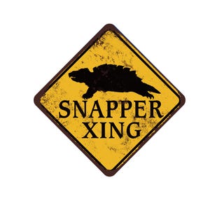 May include: A yellow diamond-shaped sign with a brown border and a distressed texture. The sign features a black silhouette of a turtle and the words "SNAPPER XING" in bold, black letters. The sign is designed to look aged.