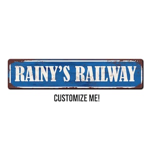 May include: A blue and brown metal sign with the text "Rainy's Railway" in white. The sign has a distressed, vintage look. The text "Customize Me!" is below the sign.